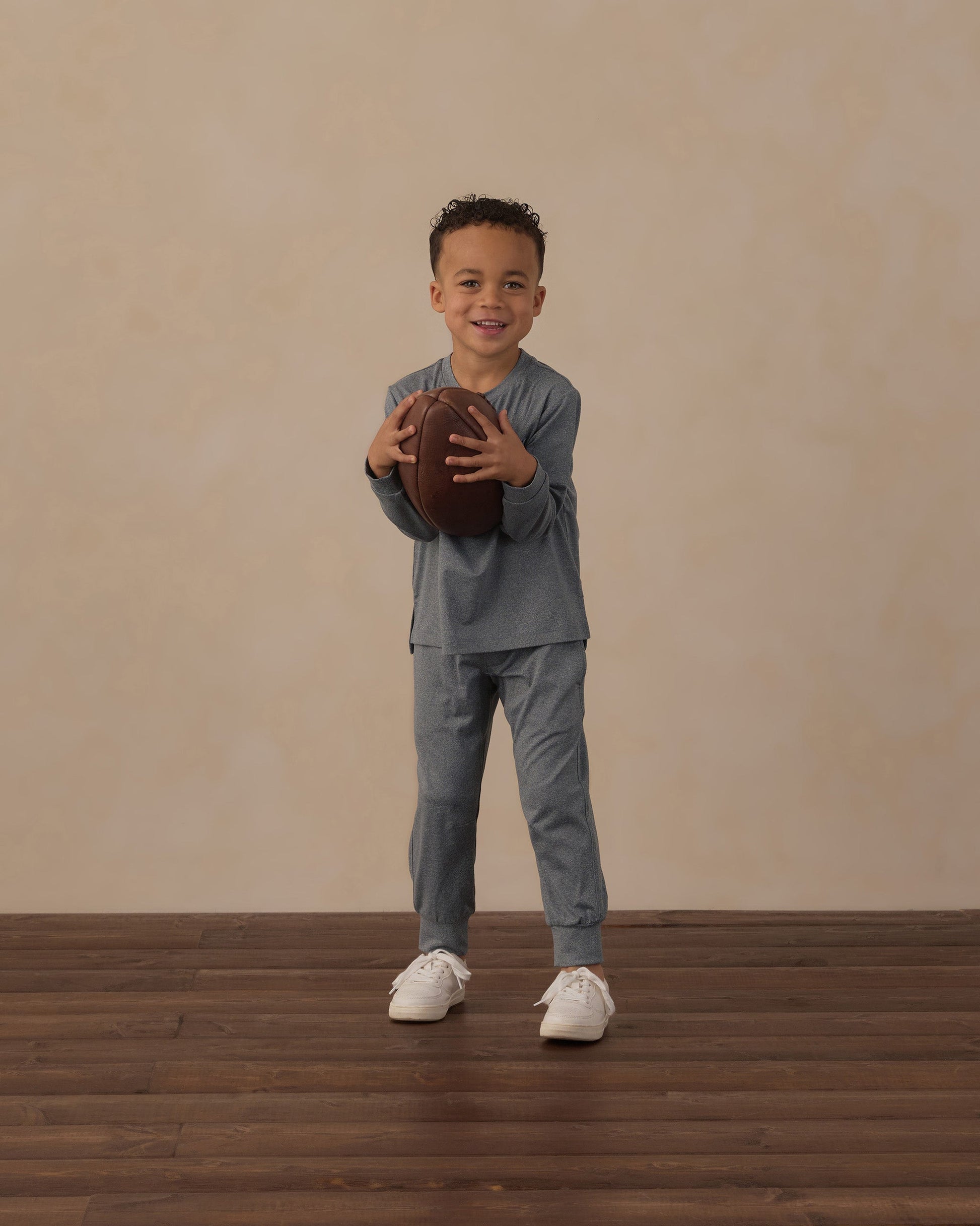 A young boy wearing Rylee & Cru Boys’ Century Tech Jogger in blue stands on a wooden floor, holding a football and smiling at the camera against a beige background.