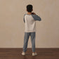 A child with curly hair stands facing away, wearing a white and blue long-sleeve shirt, Rylee & Cru Boys’ Century Tech Jogger in blue, and white sneakers on a wooden floor with a beige wall background.