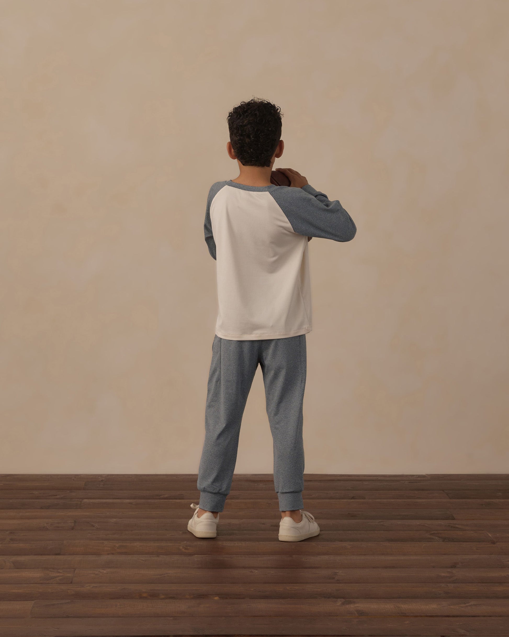 A child with curly hair stands facing away, wearing a white and blue long-sleeve shirt, Rylee & Cru Boys’ Century Tech Jogger in blue, and white sneakers on a wooden floor with a beige wall background.