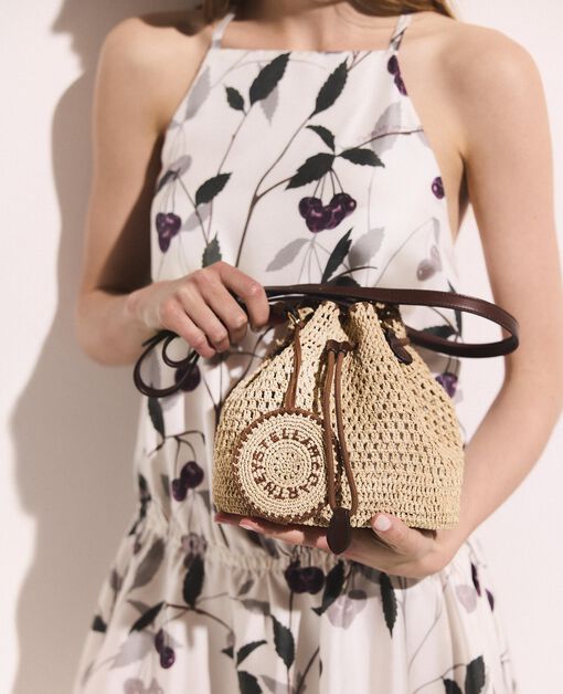 A woman in a sleeveless, floral dress holds Stella McCartney’s Falabella Crochet Raffia Bucket Bag, a small beige luxury vegan accessory with a brown leather strap and circular woven detail, echoing the brand's signature tote style.