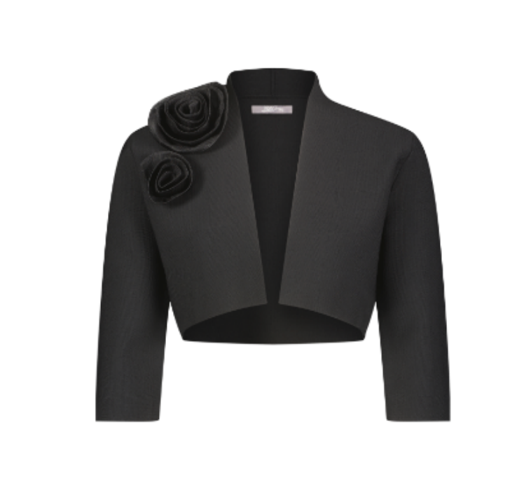 The Rose Bolero by Lela Rose is a black cropped jacket in a soft polyester-viscose blend, featuring three-quarter sleeves and two large fabric rose accents on the left shoulder. Imported for a modern, elegant look.