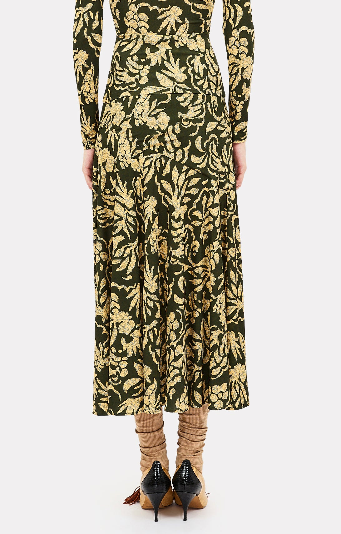 A person shown from the back against a white background wears the Ulla Johnson Fiona Jersey Skirt, which features a gold floral pattern on dark fabric that creates the illusion of a high-waisted skirt, styled with tan socks and black pointed-toe heels.