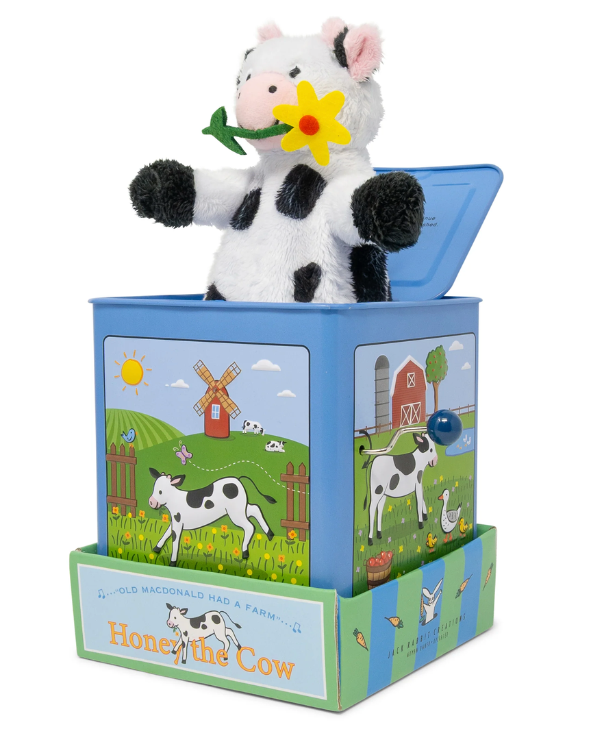 A plush cow holding a yellow flower pops out of the Jack Rabbit Creations Jack-in-the-Box, a blue box adorned with farm illustrations and the words “Honey the Cow.” This classic toy from Jack Rabbit Creations delights children with its charming plush character, bringing surprise and smiles to playtime.