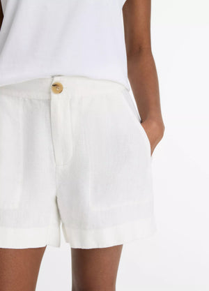 A person wearing a white short-sleeve top and vince Mid Rise Hemp Shorts in white with breathable fabric and a button closure, standing with one hand in their pocket.