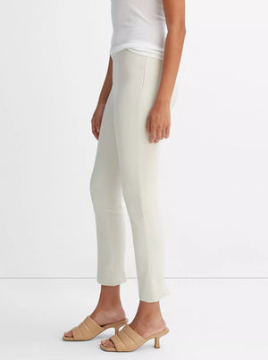 A person wearing vince’s Bobbi Mid Rise Pant in off-white, crafted from Italian cotton-blend ponte, styled with a sleeveless white top and beige heeled sandals, stands against a plain white background.