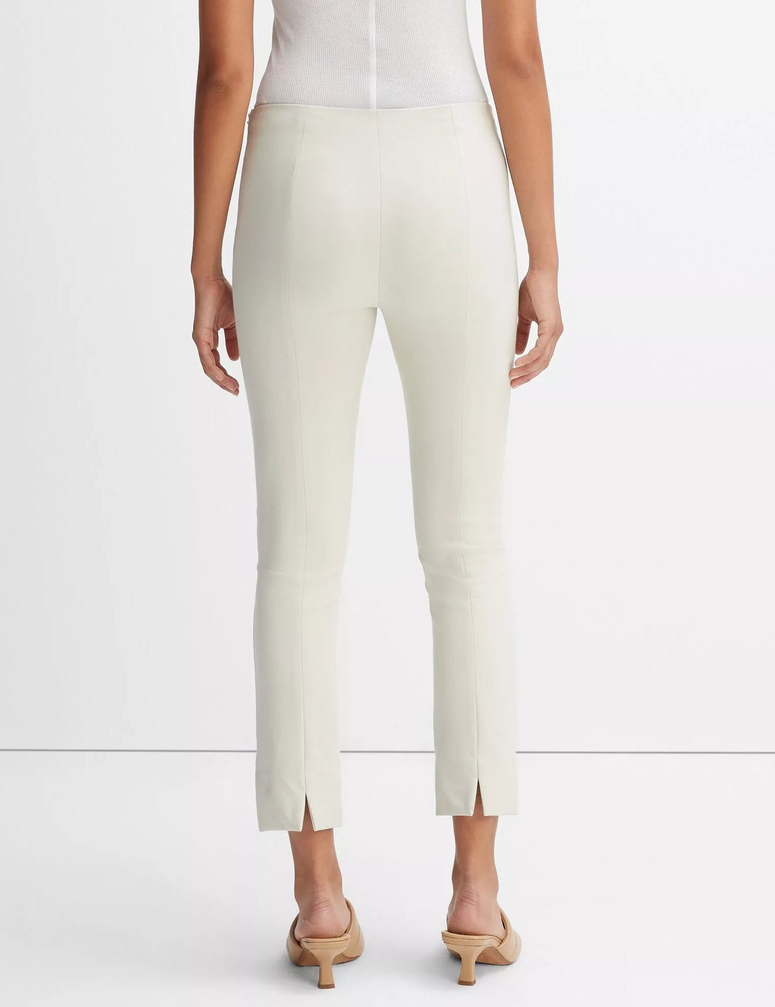 A person wearing light-colored, fitted Bobbi Mid Rise Pants by vince, crafted from an Italian cotton-blend ponte with back slits, paired with beige heeled sandals and a white sleeveless top, is shown from the back against a plain background.