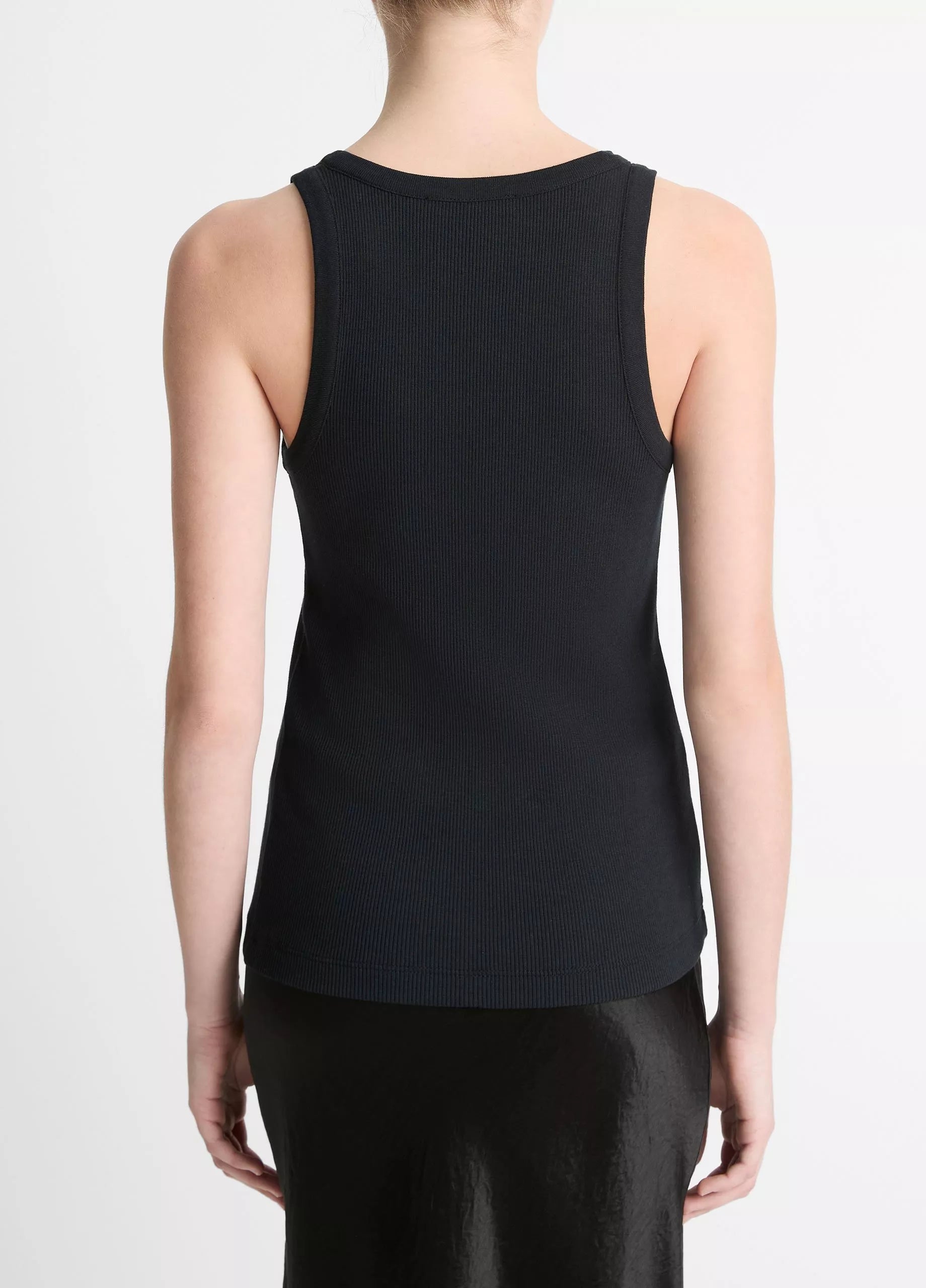 A person stands facing away in the Ribbed Scoop Neck Tank by vince— a sleeveless black tank crafted from a cotton-modal blend—paired with a black skirt, set against a plain white background.