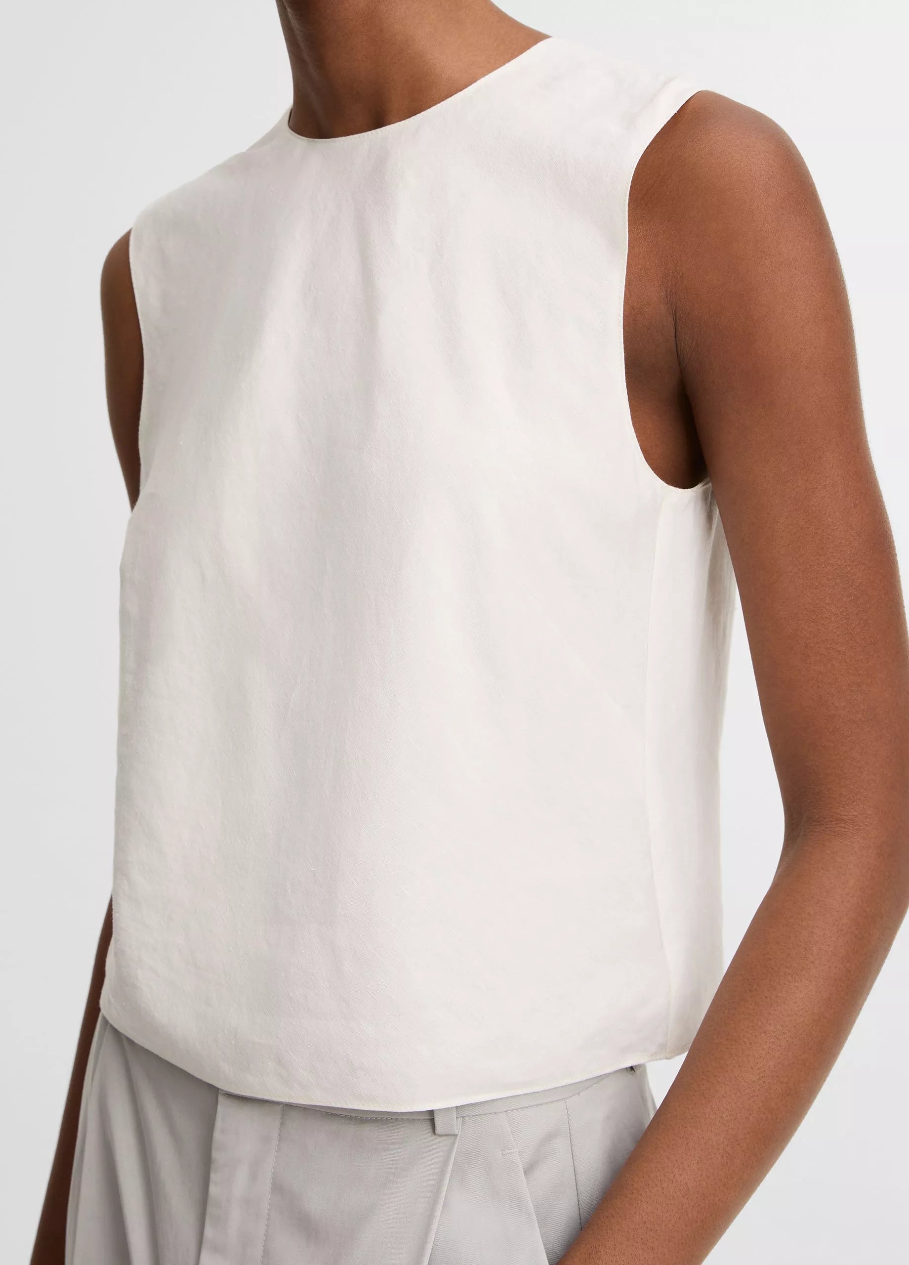 A person wears the vince Linen Crew Neck Shell Tank in white with light gray high-waisted pants, shown from shoulders to waist against a plain background. The breathable linen blend enhances comfort and offers a simple, refined look.