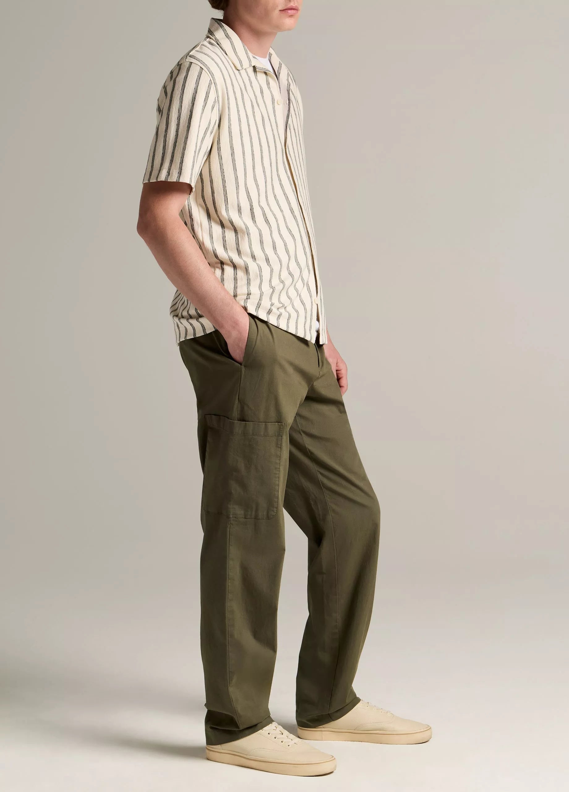 A person stands in profile wearing a short-sleeved, striped button-up shirt, the vince Owen Side Pocket Trouser in olive, and beige sneakers against a plain background.