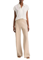 A person is wearing a short-sleeved, collared white blouse paired with the vince Colette Linen Bias Pant in beige—featuring wide legs and a hidden elastic waistband—and open-toe sandals, standing against a plain white background.