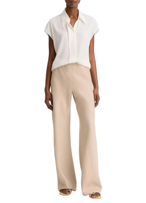 A person is wearing a short-sleeved, collared white blouse paired with the vince Colette Linen Bias Pant in beige—featuring wide legs and a hidden elastic waistband—and open-toe sandals, standing against a plain white background.