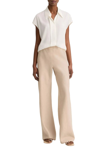 A person is wearing a short-sleeved, collared white blouse paired with the vince Colette Linen Bias Pant in beige—featuring wide legs and a hidden elastic waistband—and open-toe sandals, standing against a plain white background.