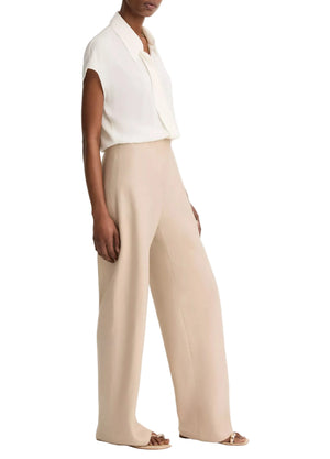 A person wears a short-sleeve white blouse tucked into the vince Colette Linen Bias Pant—high-waisted, wide-leg beige pants crafted from Italian linen and featuring a hidden elastic waistband—paired with beige heeled sandals, standing against a plain white background.