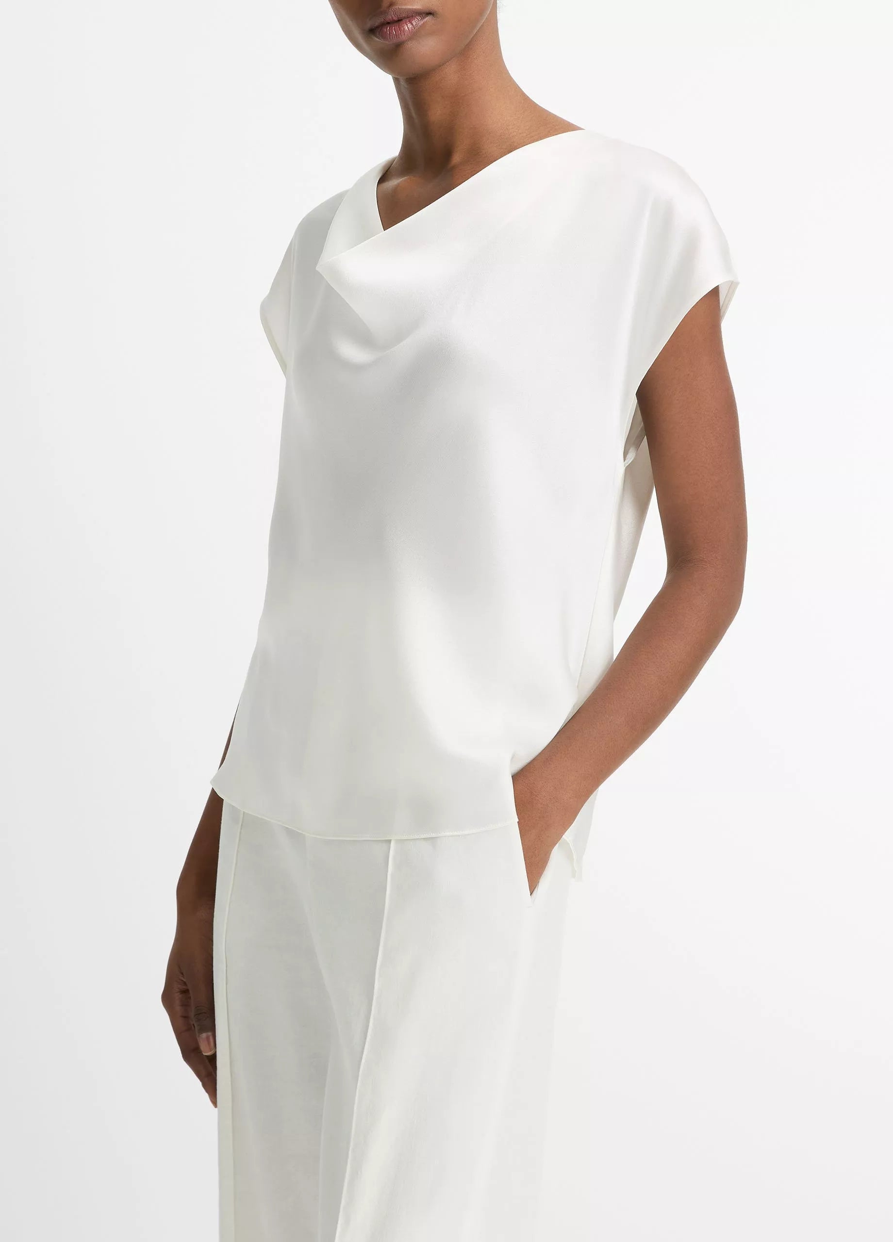A woman wearing the Vince Cowl Neck Cap Sleeve Silk Blouse, styled in a loose fit and paired with matching white trousers, stands against a plain white background.