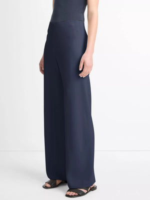 A person wearing a sleeveless black top, dark blue Colette High Waist Pant by vince with an elastic waistband, and black sandals stands against a plain white background.