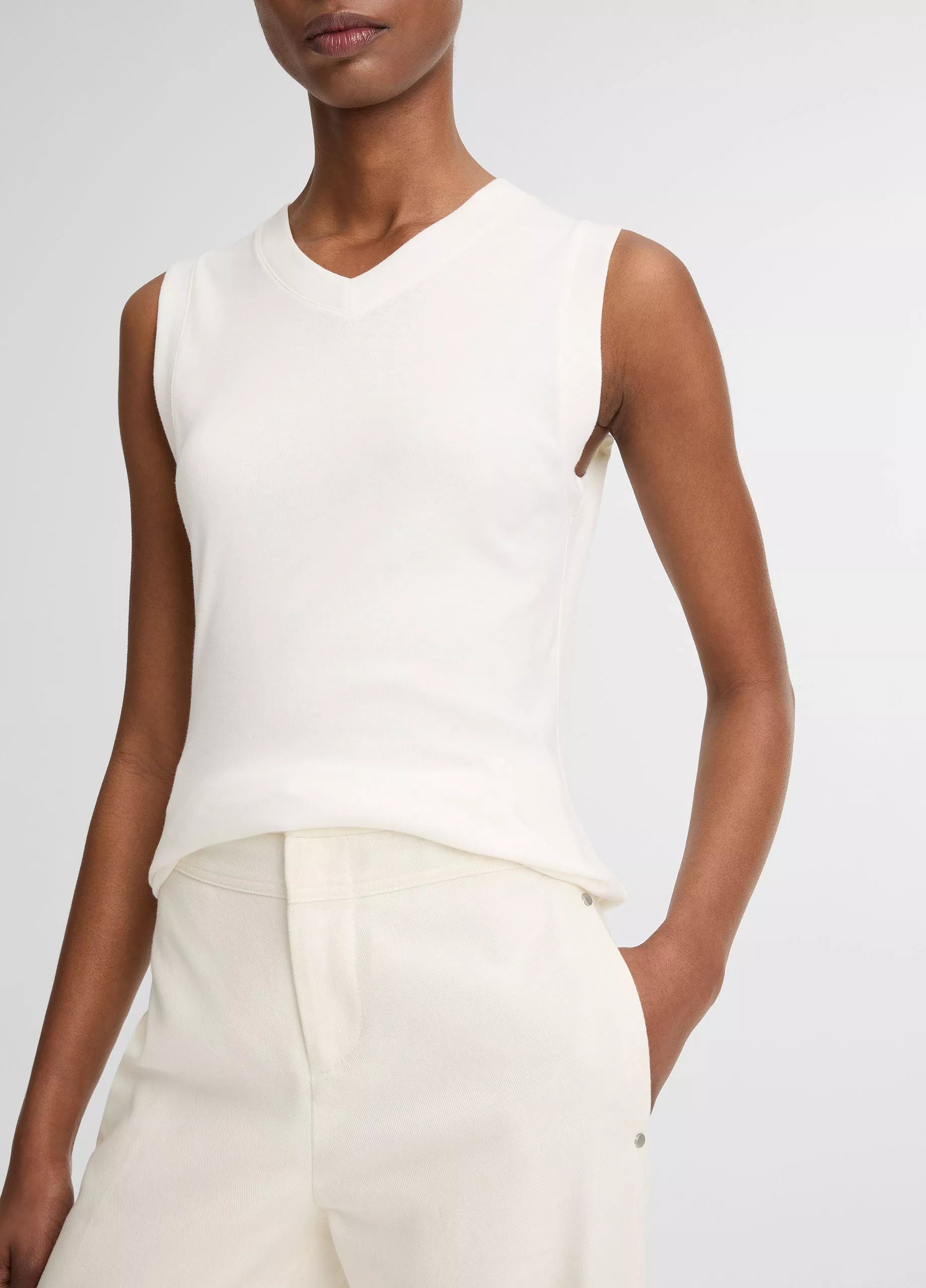 A person wearing the Vince High V-Neck Shell Tank in white and high-waisted white pants, with one hand in their pocket, stands against a plain background.