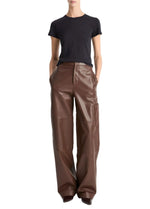 A person wearing the vince Essential Crew black short-sleeve t-shirt, crafted from soft Peruvian Pima cotton, tucked into high-waisted, wide-leg brown leather pants with hands in pockets, stands against a plain white background.