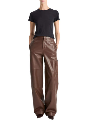 A person wearing the vince Essential Crew black short-sleeve t-shirt, crafted from soft Peruvian Pima cotton, tucked into high-waisted, wide-leg brown leather pants with hands in pockets, stands against a plain white background.