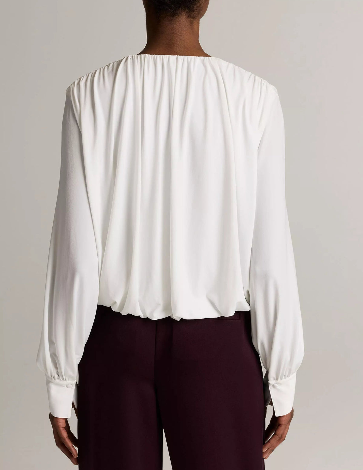 A person is shown from behind wearing the Vince Matte Jersey V-Neck Bubble Top—a long-sleeve, white blouse with gathered details—paired with dark high-waisted pants, standing against a plain background for a minimal yet subtly sophisticated look.