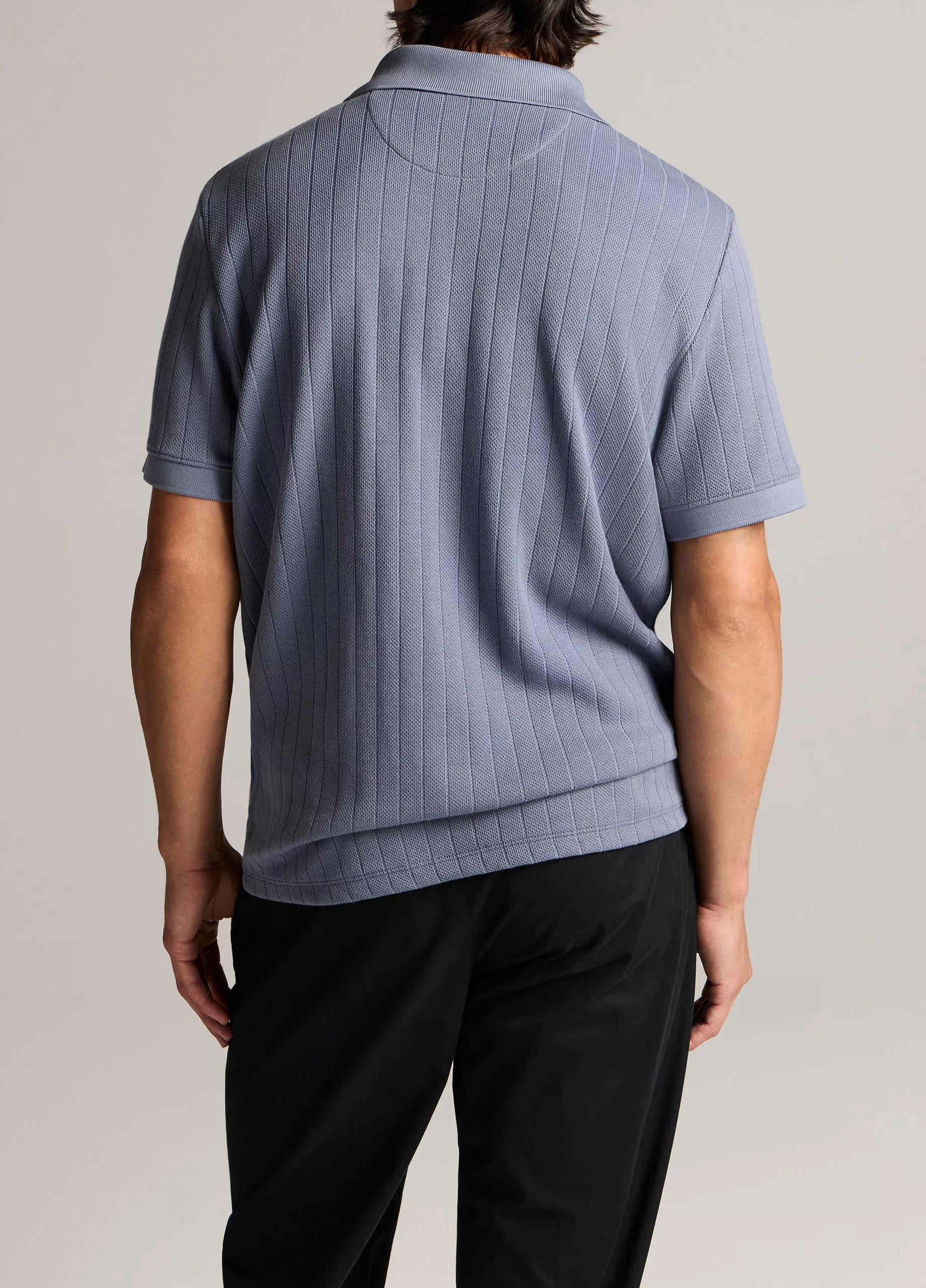 A person stands facing away, wearing the vince Drop Needle Stripe SS Polo in light purple and black pants against a neutral background.