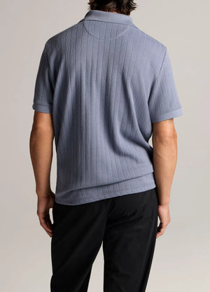 A person stands facing away, wearing the vince Drop Needle Stripe SS Polo in light purple and black pants against a neutral background.