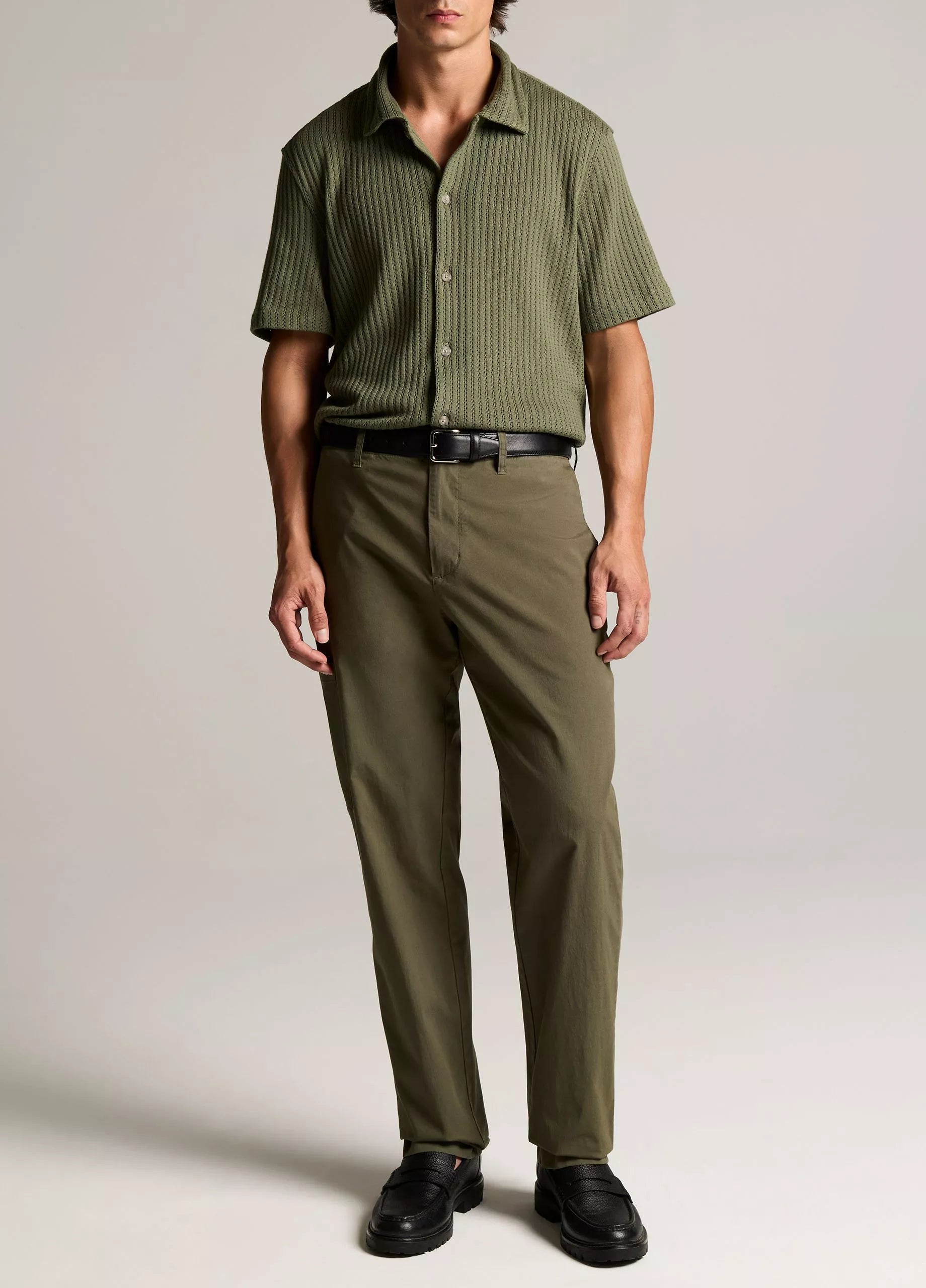 A man wearing the vince Pointelle Short Sleeve Button Down in olive, paired with matching olive trousers, a black belt, and black shoes, stands against a plain background.