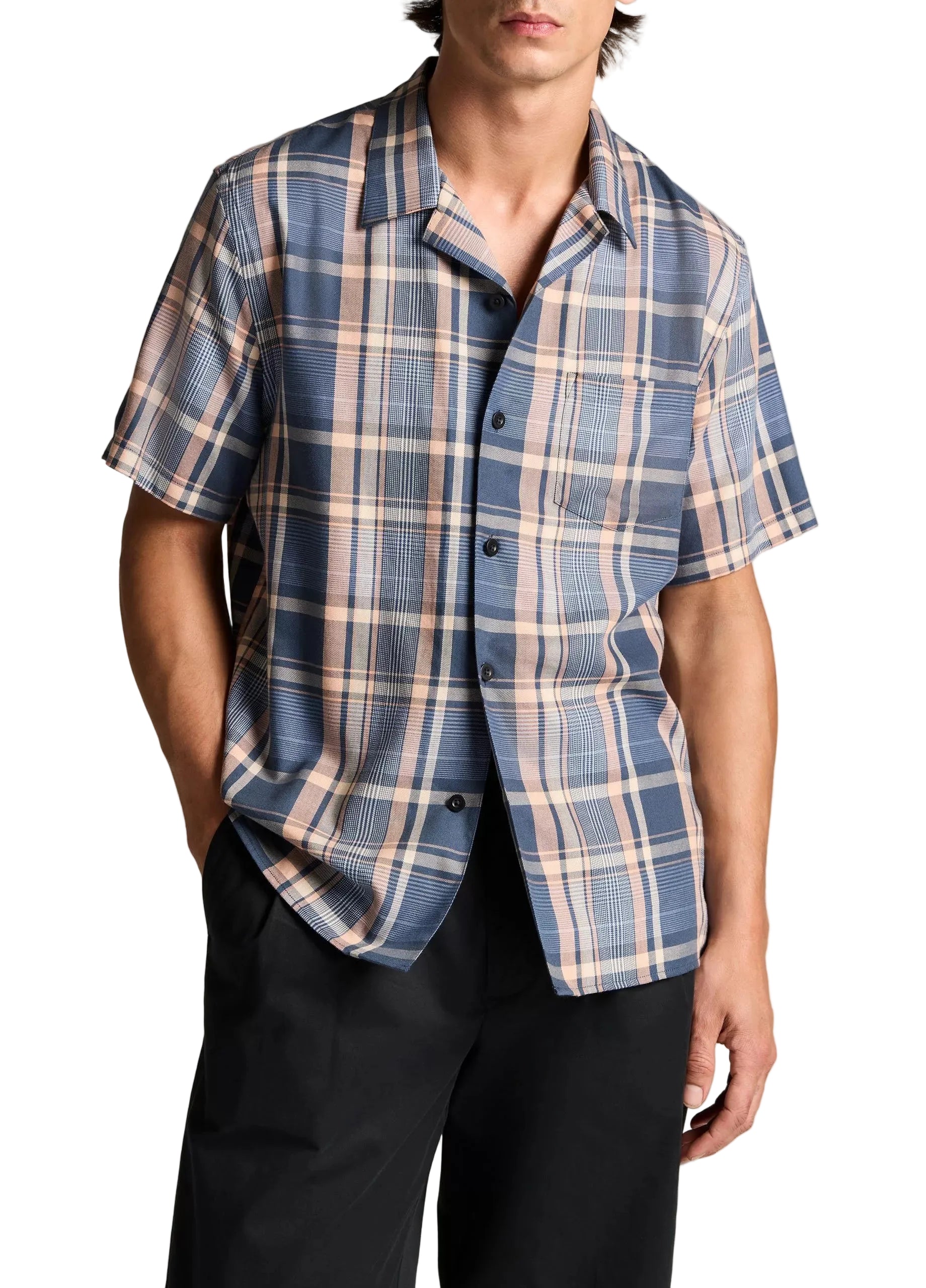 A person wearing the vince Prine Plaid Short Sleeve Cabana—a classic blue and beige plaid shirt crafted from comfortable cotton-blend twill—paired with black pants and standing with one hand in a pocket.