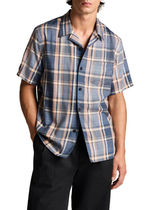 A person wearing the vince Prine Plaid Short Sleeve Cabana—a classic blue and beige plaid shirt crafted from comfortable cotton-blend twill—paired with black pants and standing with one hand in a pocket.