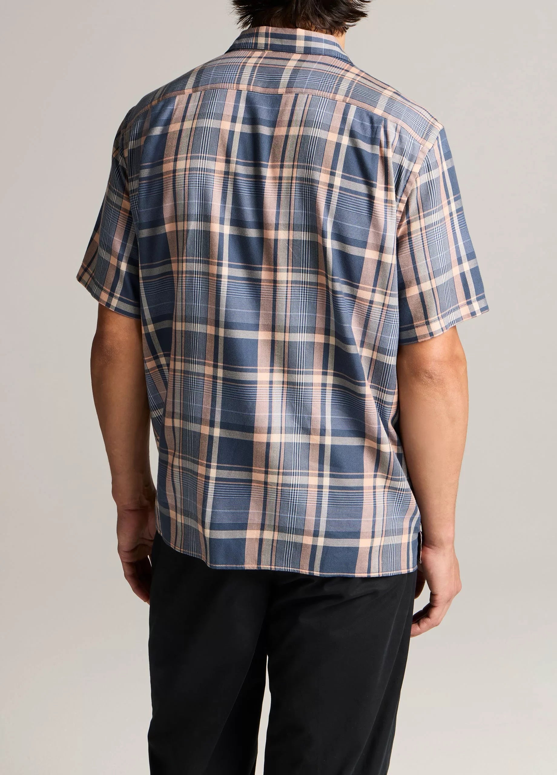 A person is seen from the back wearing the vince Prine Plaid Short Sleeve Cabana shirt in blue, beige, and pink cotton-blend twill with black pants, set against a plain background.