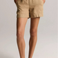 A person wearing the Vince Mid Rise Hemp Short in tan, styled with a relaxed silhouette, paired with a white short-sleeve shirt and brown sandals, stands with hands in pockets against a plain background.
