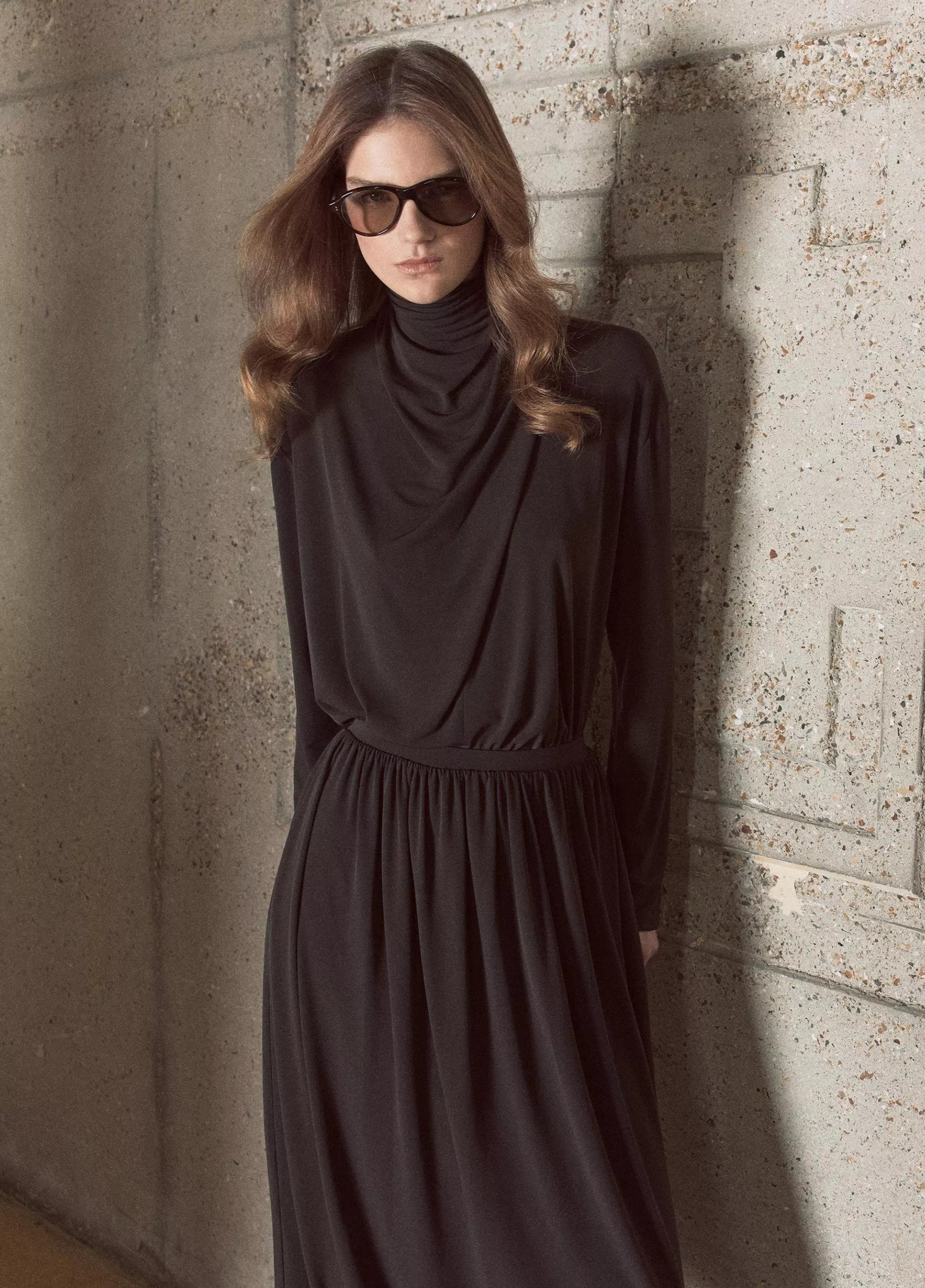 A person wearing sunglasses and a long sleeve draped cowl blouse by vince stands against a textured concrete wall.