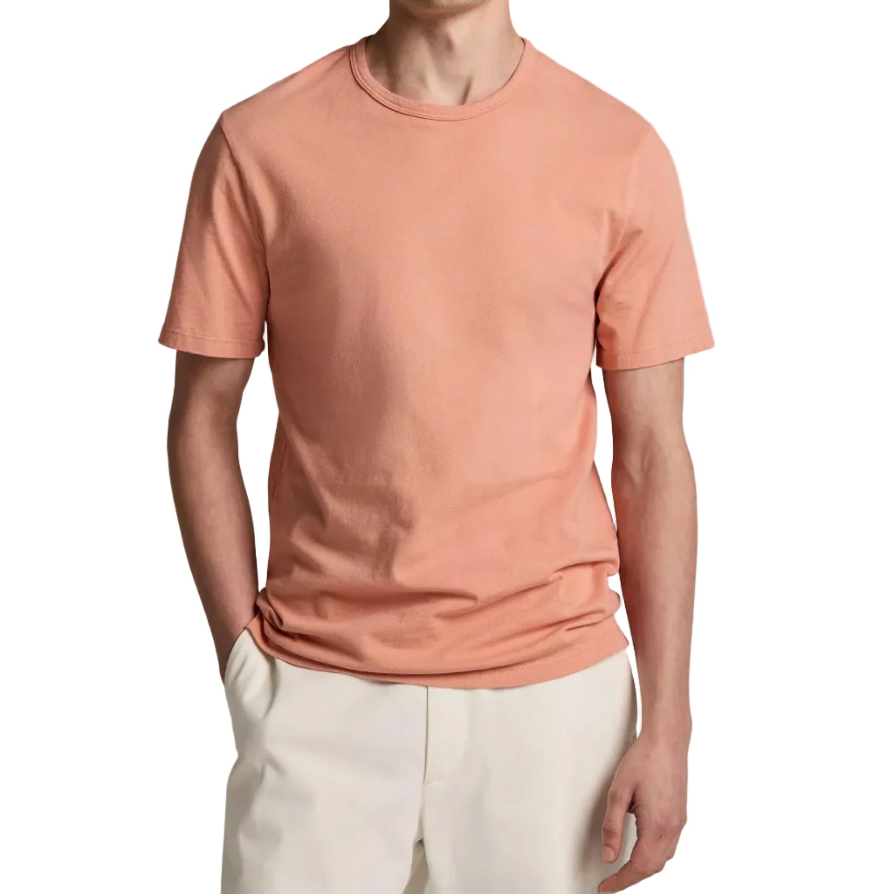 A person wearing the Vince Garment Dye S/S Crew, an ultra-soft, peach-colored cotton jersey short-sleeve t-shirt, paired with light beige pants and standing with one hand in their pocket against a white background.