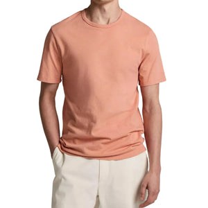 A person wearing the Vince Garment Dye S/S Crew, an ultra-soft, peach-colored cotton jersey short-sleeve t-shirt, paired with light beige pants and standing with one hand in their pocket against a white background.