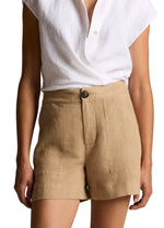 A person wearing a white short-sleeve button-down shirt tucked into the vince Mid Rise Hemp Short, a pair of high-waisted, tan breathable shorts with a relaxed silhouette, complete with front button and pocket detailing.