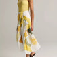 A person wears a sleeveless yellow top paired with the vince Painted Abstract Draped Skirt—a white midi twill skirt adorned with large yellow and green floral motifs and crafted from LENZING™ ECOVERO™ viscose—styled with brown sandals, standing against a plain background.