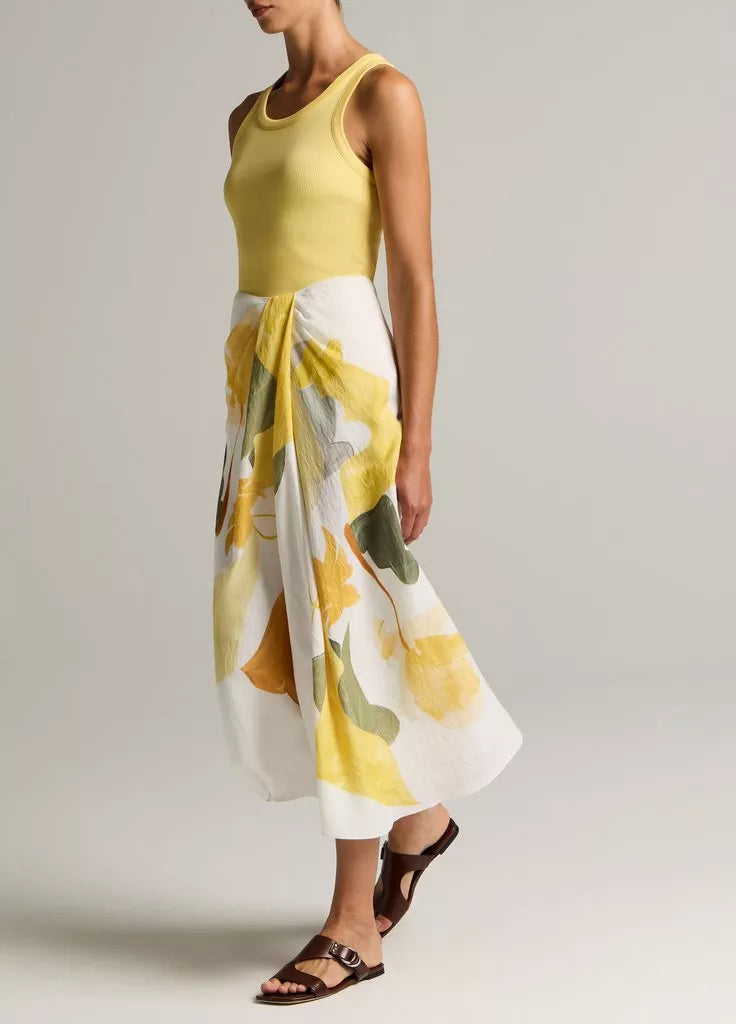 A person wears a sleeveless yellow top paired with the vince Painted Abstract Draped Skirt—a white midi twill skirt adorned with large yellow and green floral motifs and crafted from LENZING™ ECOVERO™ viscose—styled with brown sandals, standing against a plain background.