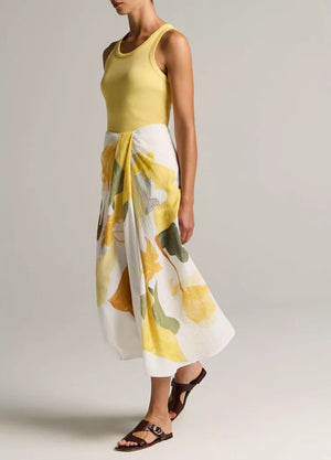 A person wears a sleeveless yellow top paired with the vince Painted Abstract Draped Skirt—a white midi twill skirt adorned with large yellow and green floral motifs and crafted from LENZING™ ECOVERO™ viscose—styled with brown sandals, standing against a plain background.