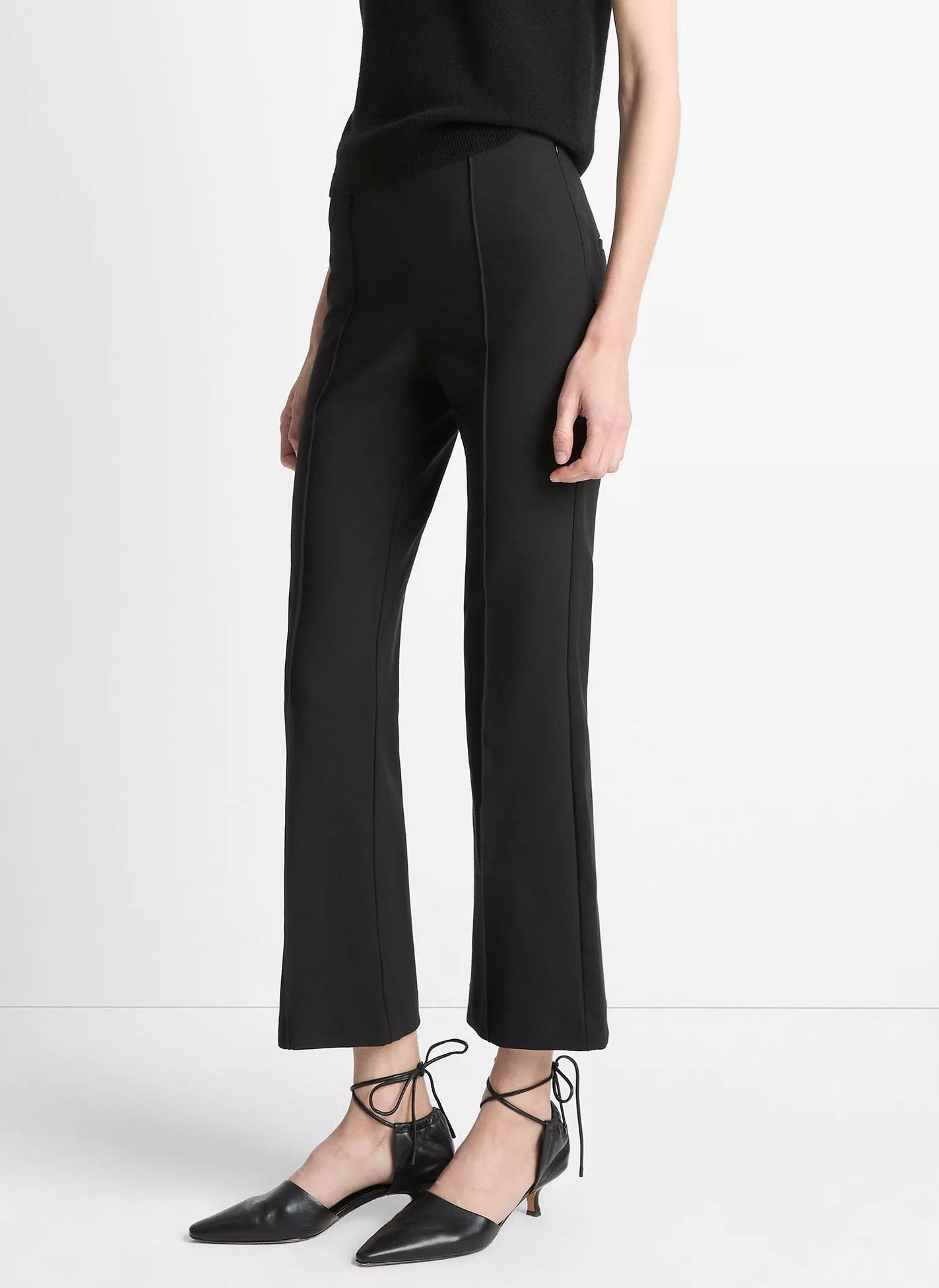 A person wearing Vince’s Niki Mid Rise Crop Flare Pant in black Italian cotton-blend, featuring pintuck legs, paired with black pointed-toe ankle-strap shoes and standing against a plain white background.