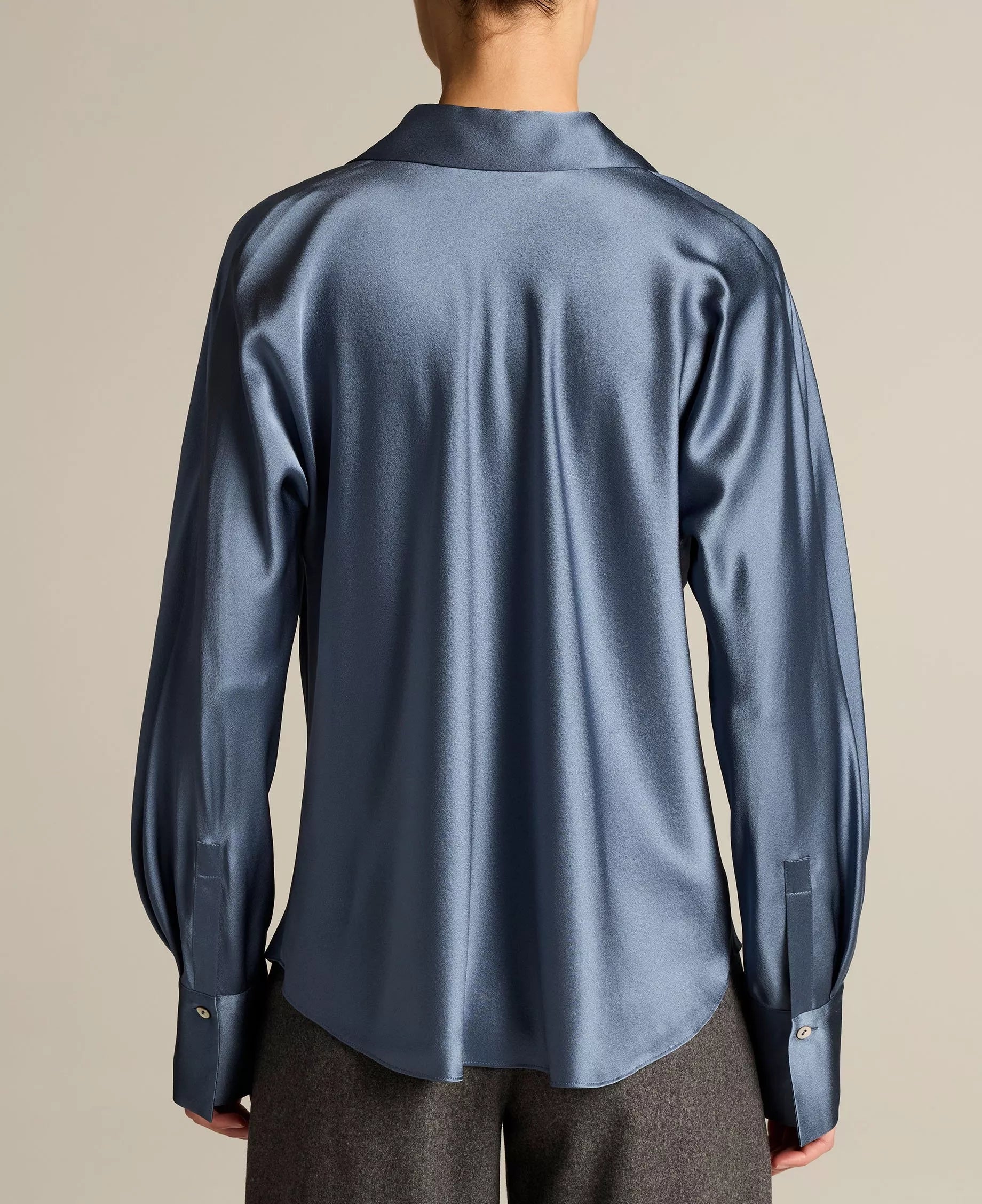 A person is seen from the back wearing the Vince Bias Long Sleeve Blouse in blue, paired with gray pants, standing against a plain light background.