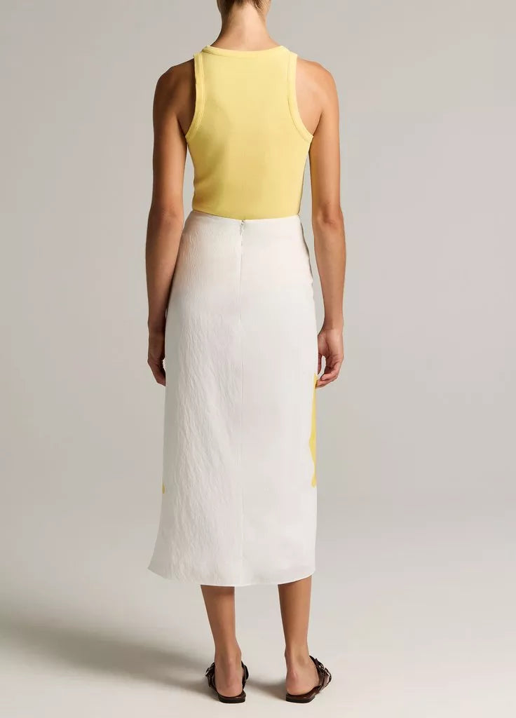 A person stands barefoot, facing away in a plain studio, wearing a yellow sleeveless top paired with the Painted Abstract Draped Skirt by vince—a white midi twill skirt crafted from LENZING™ ECOVERO™ viscose that lends an elegant touch to the simple ensemble.