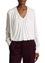 A person wears the Vince Matte Jersey V-Neck Bubble Top in white, paired with burgundy pants and stands with one hand in their pocket, capturing a minimal interpretation of effortless style.