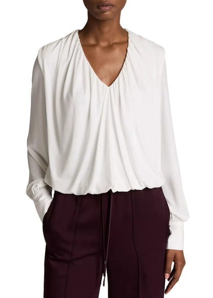 A person wears the Vince Matte Jersey V-Neck Bubble Top in white, paired with burgundy pants and stands with one hand in their pocket, capturing a minimal interpretation of effortless style.