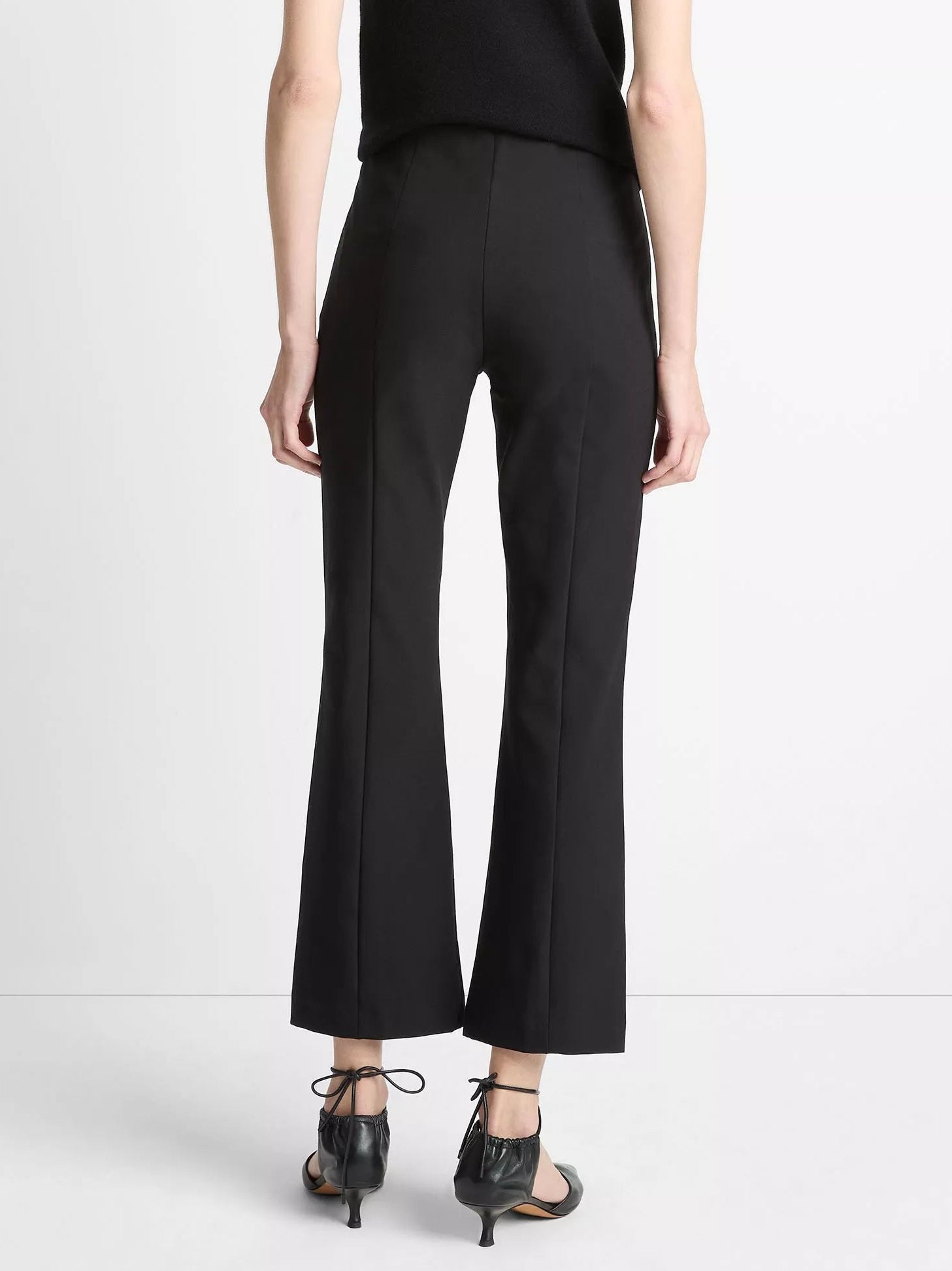A person facing away wears black vince Niki Mid Rise Crop Flare Pants with pintuck legs and black heeled sandals with ankle ties, standing against a white background.