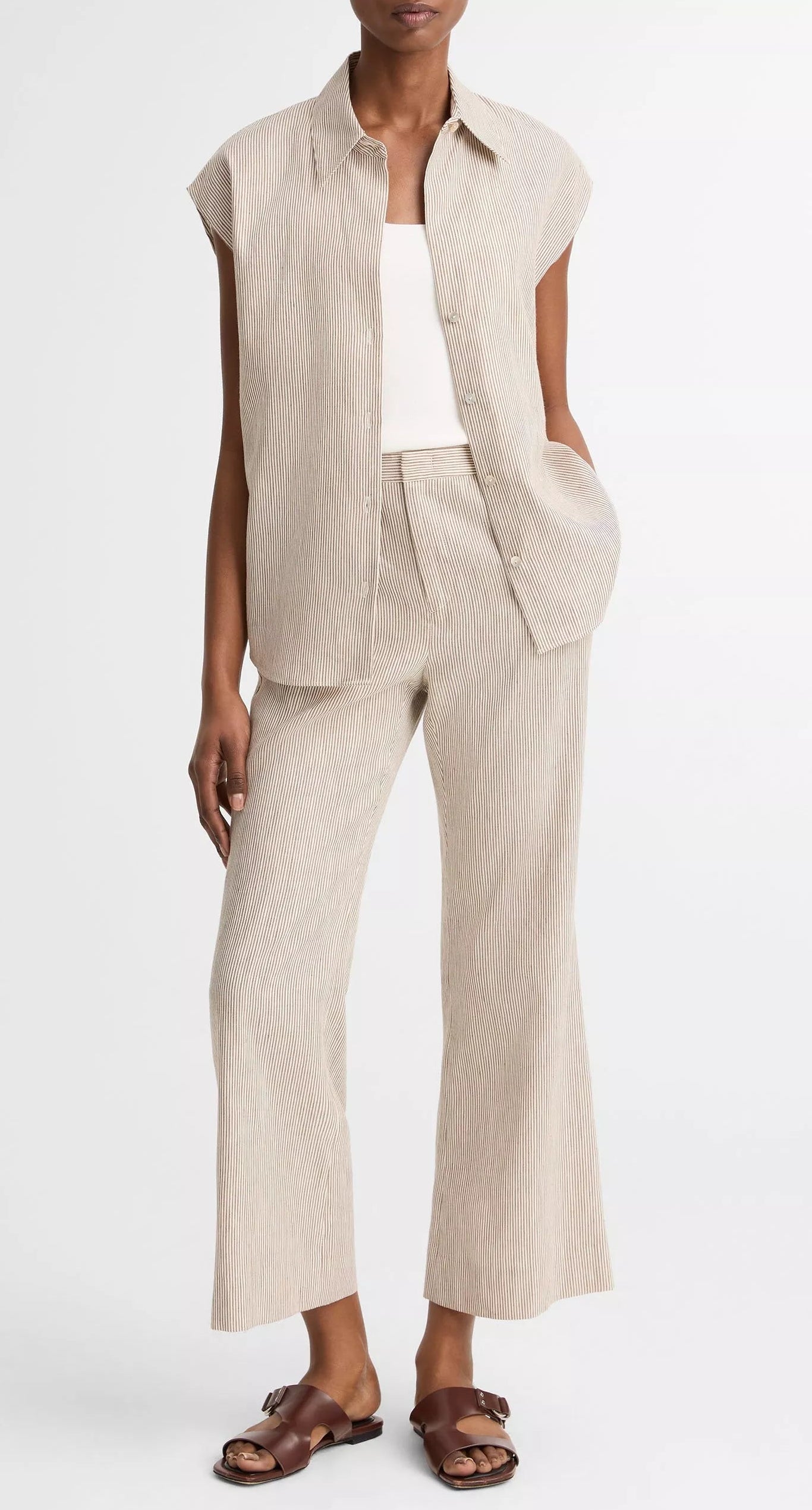 A person stands against a plain background, wearing a beige pinstriped short-sleeve shirt, white top, brown sandals, and vince Low Rise Ticking Striped Crop Flare Trousers.