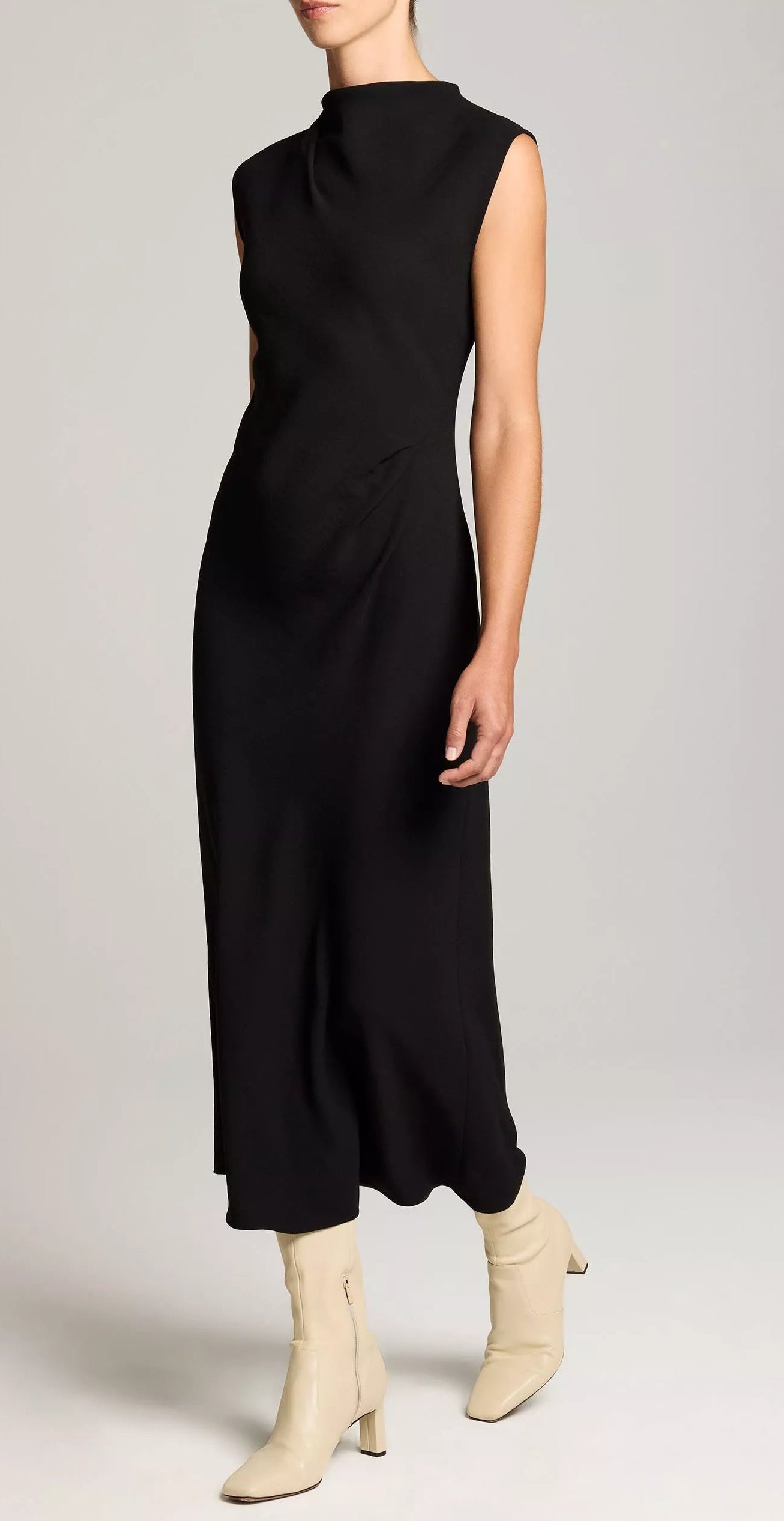 A person is wearing the vince Draped Funnel Neck Dress in black matte crepe, featuring a sleeveless design and pleated details, paired with light beige ankle boots, standing against a plain light background.