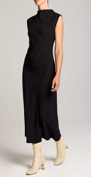 A person is wearing the vince Draped Funnel Neck Dress in black matte crepe, featuring a sleeveless design and pleated details, paired with light beige ankle boots, standing against a plain light background.