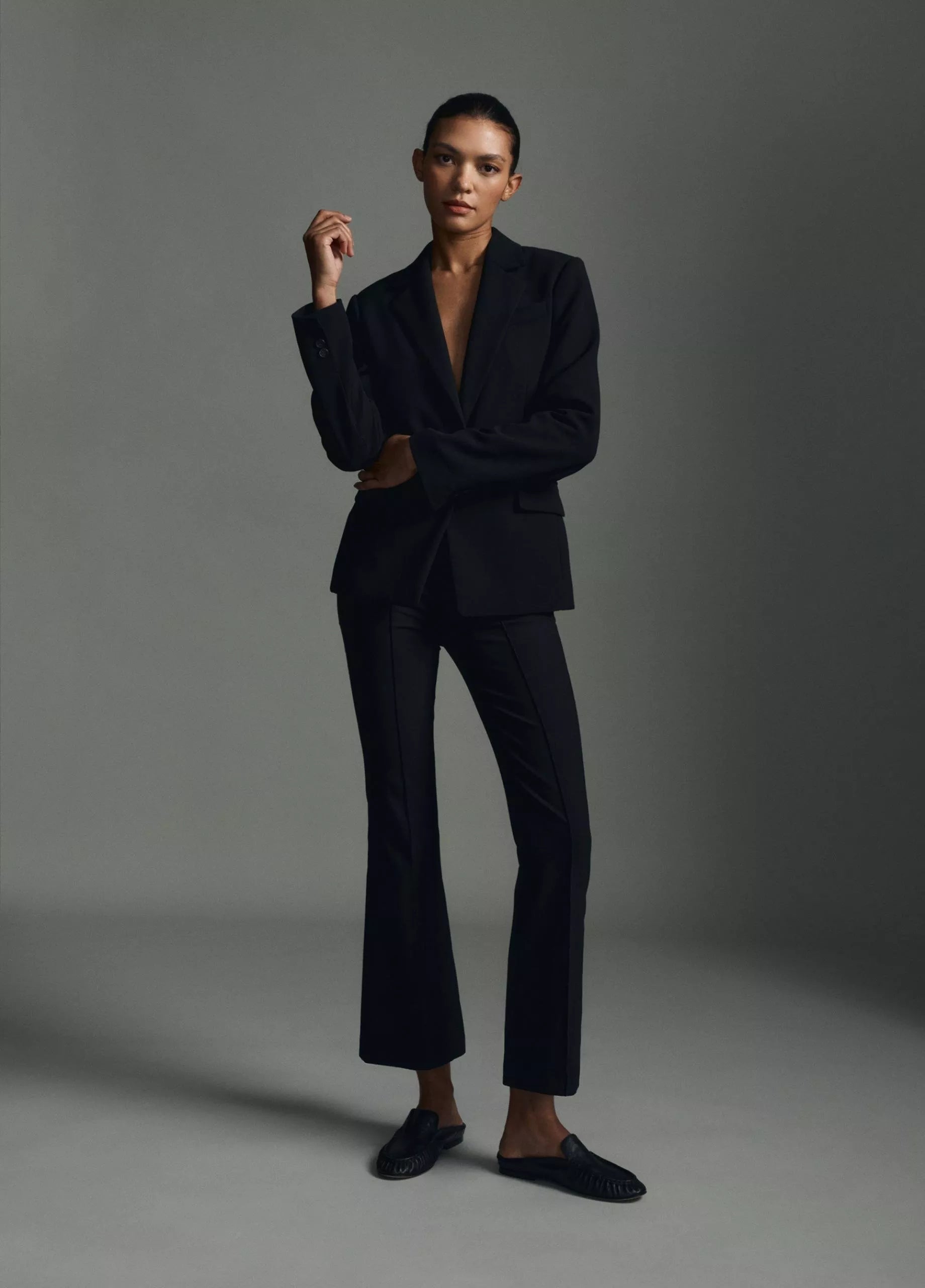 A person stands against a plain background wearing the vince Niki Mid Rise Crop Flare Pant in black Italian cotton-blend fabric, styled without a shirt and paired with sleek black flat shoes.