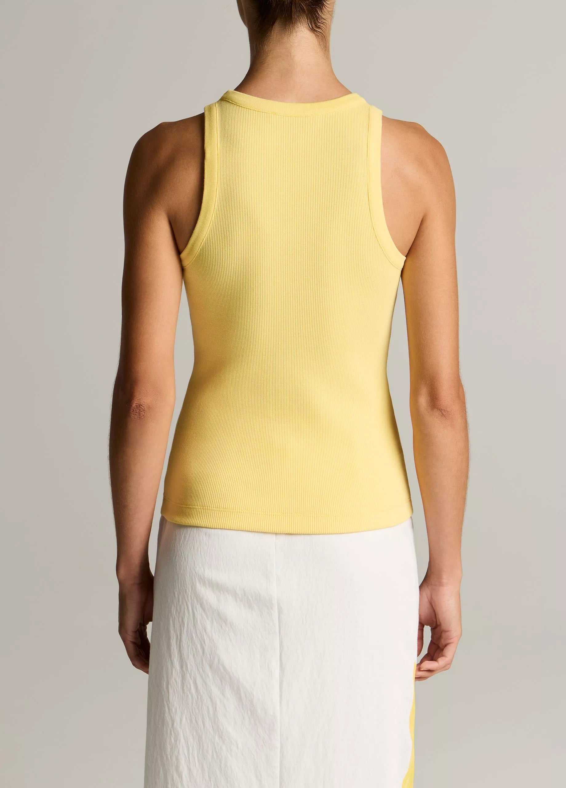 A person stands with their back to the camera, wearing the vince Ribbed Scoop Neck Tank in yellow—a finely ribbed, sleeveless cotton-modal blend top—paired with a white skirt featuring a yellow stripe, set against a plain background.
