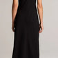 A person stands facing away against a plain light background, wearing the Vince Draped Funnel Neck Dress in black matte crepe and tall cream-colored boots.