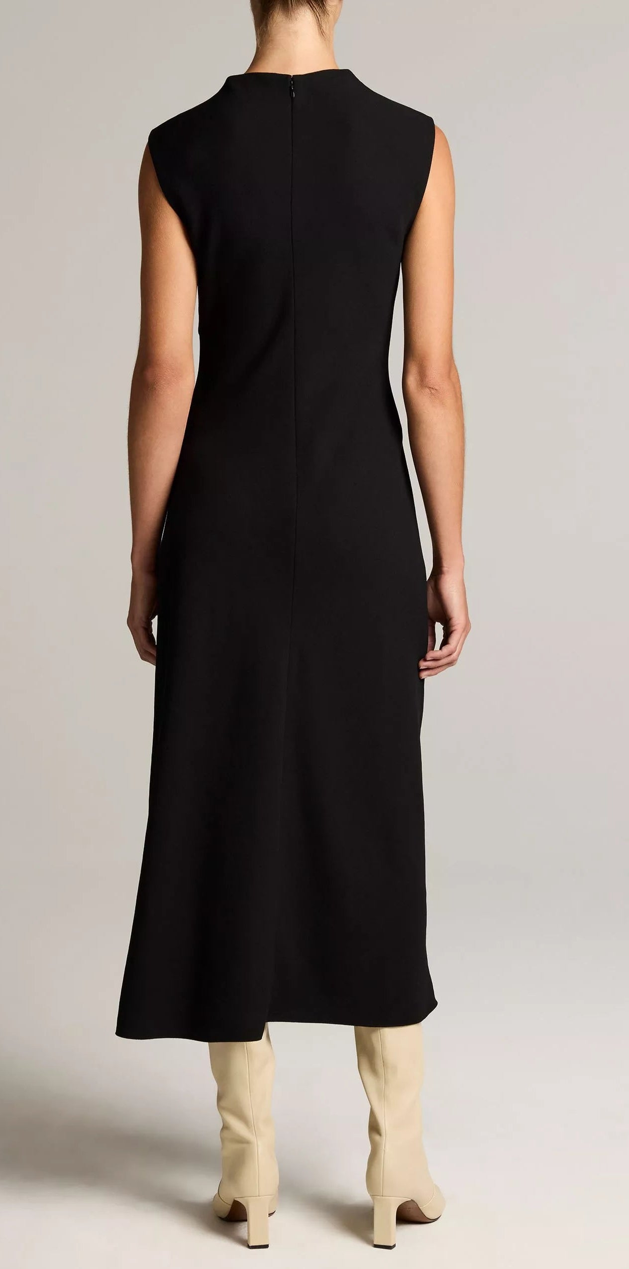 A person stands facing away against a plain light background, wearing the Vince Draped Funnel Neck Dress in black matte crepe and tall cream-colored boots.